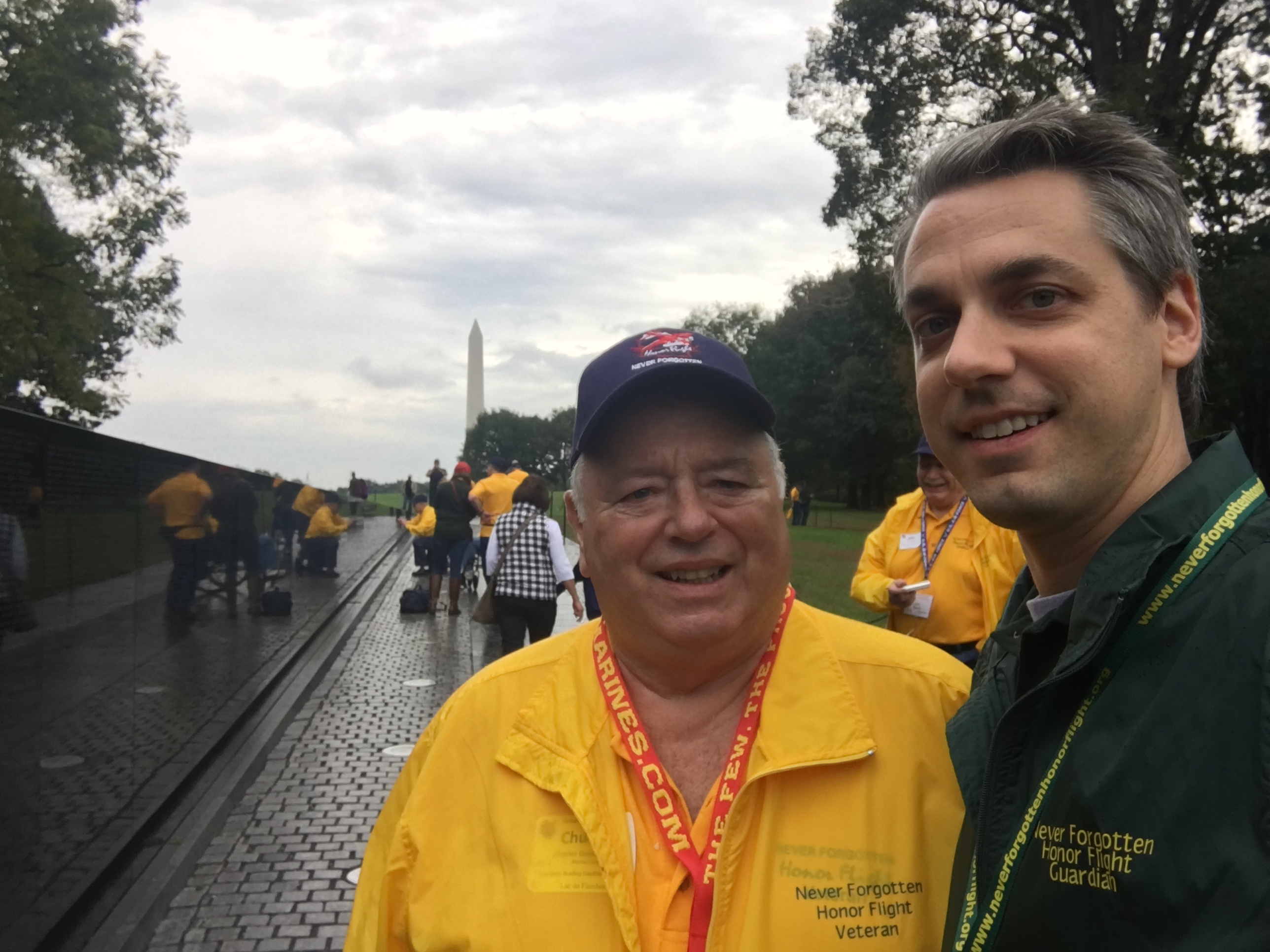 My dad and me at the Vietnam Veterans Memorial in Washington DC, on the Never Forgotten Honor Flight. He is wearing a yellow Honor Flight veteran jacket. I am wearing a green guardian jacket. The Washington Monument is visible in the distance.